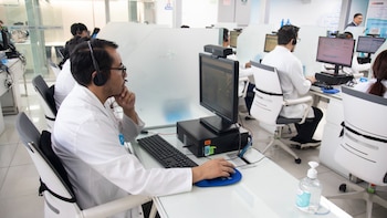 Múltiples personas con batas blancas y auriculares sentadas en escritorios individuales con computadoras en un centro de atención