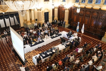 Interior de un gran salón con personas sentadas en mesas largas y sillas, una pantalla de proyección, dos banderas (una de Guatemala), candelabros y pilares decorados