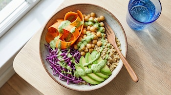 Vista aérea de un bowl lleno de quinoa, garbanzos, zanahoria en tiras, repollo morado y aguacate en rodajas con aderezo verde, junto a un vaso de agua azul.