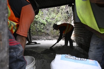 Varias personas, incluyendo hombres, mujeres y niños, sentadas en mesas largas comiendo de platos con comida variada y bebidas en vasos de plástico