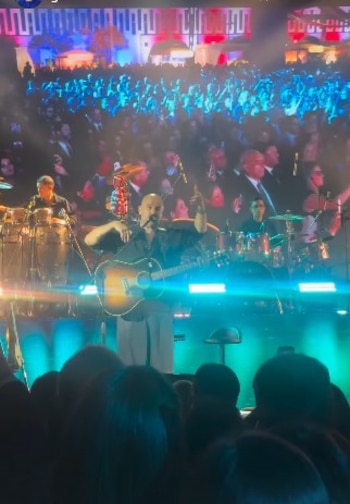 Cantante con guitarra acústica en un escenario con luces azules. Detrás hay otros músicos y una pantalla con un edificio y una multitud. Público en primer plano