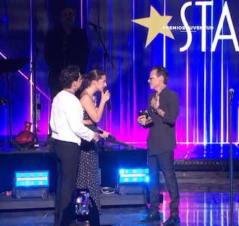 Ángela Aguilar habla por micrófono junto a Christian Nodal ante Marc Anthony, quien sostiene un trofeo en un escenario con fondo azul
