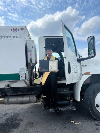 Un hombre con toga, birrete y estola de graduación sostiene su título junto a un camión municipal blanco con la puerta abierta
