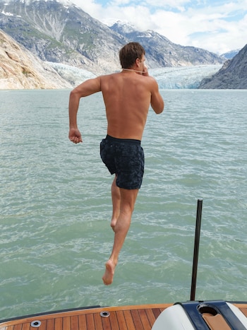 Hombre salta al agua, sin camiseta y con bañador oscuro. Al fondo, lago, montañas rocosas, glaciares y cielo con nubes. Cubierta de barco en primer plano