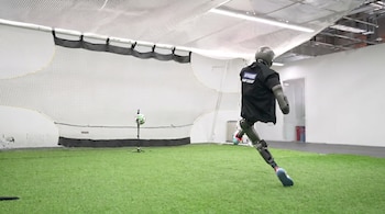 Un robot con chaleco negro y piernas articuladas corre en un campo de césped artificial hacia un balón de fútbol, con una portería al fondo.