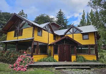 Casa de madera de dos pisos con paredes amarillas, ventanales grandes, un porche de madera y un arbusto con flores rosadas al frente
