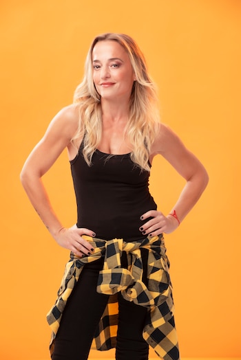 Maren Morris de pie, cabello rubio, sonriendo. Viste top y pantalones negros, camisa a cuadros amarillos y negros atada a la cintura, frente a un fondo naranja