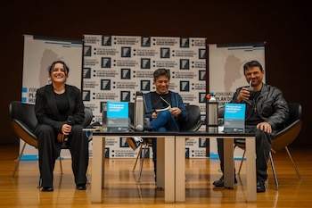 Tres personas sentadas en un escenario frente a una mesa con termos, libros y micrófonos, delante de paneles con inscripciones