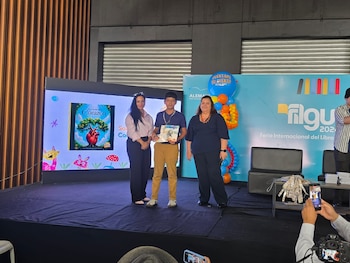 Escenario con tres personas. Un joven al centro sostiene un libro. Dos mujeres lo acompañan. Fondo con pantalla, cartel FILGU 2023 y globos decorativos