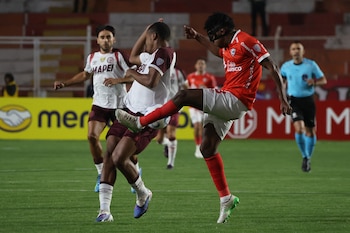 Dos jugadores de fútbol disputan un balón en un campo de césped. Uno lleva camiseta roja y máscara negra. Otro usa camiseta blanca y burdeos. Hay un árbitro y gradas