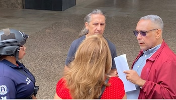 Cuatro personas. Agente de seguridad con casco y uniforme oscuro. Hombre con pelo largo gris. Mujer con pelo rubio. Hombre con gafas de sol y chaqueta roja sostiene papeles
