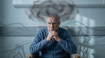 Hombre mayor de cabello gris en suéter azul, sentado en silla con manos entrelazadas y nube oscura sobre su cabeza, en un fondo azul-gris con ondas sutiles.