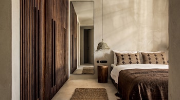 Vista de un dormitorio moderno con cama hecha, mesita de noche de tronco de madera, pared de listones de madera y un gran espejo que refleja un pasillo.