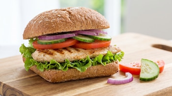 Sándwich de pollo a la parrilla con pan integral, lechuga, tomate, pepino, cebolla morada sobre tabla de madera. Rodajas de vegetales al lado.