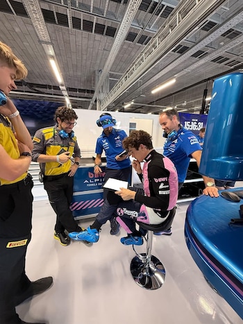 Cinco hombres en un garaje. Un piloto con traje rosa y negro sentado en un taburete, mirando una tableta. Cuatro hombres de pie a su alrededor con uniformes de equipo y auriculares. Una sección de coche azul es visible a la derecha