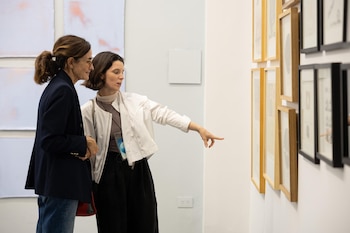 Dos mujeres de pie miran cuadros enmarcados en una pared blanca y una de ellas señala con el dedo una de las obras