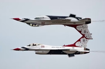 Dos aviones de combate F-16 blancos, rojos y azules de la Fuerza Aérea de EE.UU. volando en formación cerrada; uno invertido sobre el otro contra un cielo gris claro