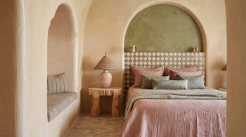 Dormitorio con paredes color arena, cama con ropa de cama rosa y cojines verdes, cabecero de azulejos, mesa de madera y lámpara tejida. Un banco acolchado se integra en una de las alcobas arqueadas.