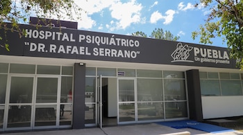 Fachada de hospital psiquiátrico con cartel que dice 'Dr. Rafael Serrano' y 'Puebla', entrada de vidrio, rampa azul y vegetación.