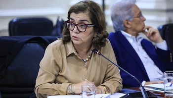 Una mujer con anteojos de marco rojo y camisa beige habla frente a un micrófono en una mesa de reuniones junto a un hombre