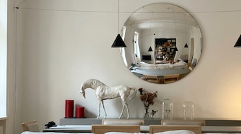 Interior con un gran espejo redondo que refleja un salón. En primer plano, una escultura de caballo blanco, velas rojas y flores secas sobre un mueble.