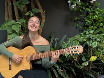 Una persona con cabello largo toca una guitarra acústica con los ojos cerrados frente a un micrófono en un escenario iluminado