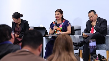 Clara Brugada Molina, con vestido azul bordado, habla por micrófono en un panel junto a otras dos personas y una audiencia; un perro descansa en el suelo