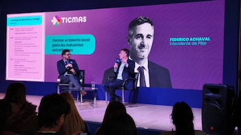 Federico Achaval, en traje oscuro, sentado en un escenario con un micrófono en mano, hablando frente a una pantalla morada que muestra su retrato ampliado y el logo de TICMAS