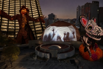 Plaza urbana con un espantapájaros gigante, una cúpula con sombras de manos proyectadas y una escultura de perro decorada como calavera.