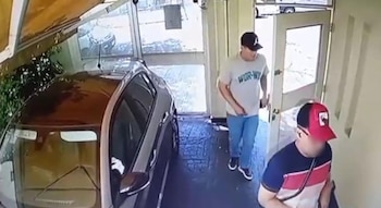Una cámara de seguridad registra a dos hombres que entran por una puerta blanca hacia un garaje donde hay un vehículo estacionado