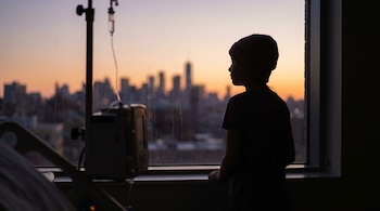 Silueta de una niña con gorro, de perfil, mirando por la ventana de un hospital al atardecer, con una ciudad al fondo. Se ven una bomba de infusión y un soporte de goteo.
