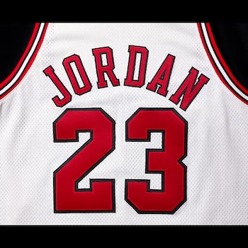 Camiseta blanca de baloncesto con el nombre Jordan y el número 23 en rojo con contorno negro. Los hombros tienen bordes negros, rojos y blancos