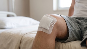 Primer plano de la rodilla de una persona sentada en una cama, cubierta con un parche o vendaje de gasa adhesivo blanco