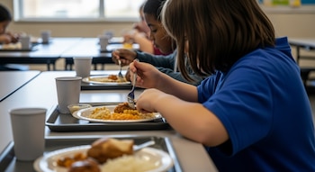 Niños sentados en mesas de cafetería escolar comen de platos blancos con tenedores. Se observan bandejas grises, comida variada y vasos de papel.