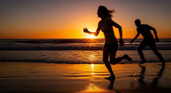 Siluetas de una mujer y un hombre corriendo en una playa con olas, reflejos del sol naranja sobre el agua y el cielo al atardecer.