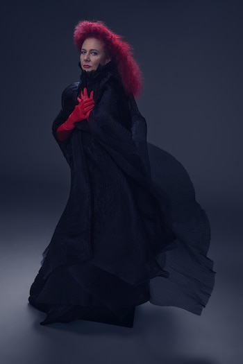 Renata Schussheim con cabello rojo, vestido largo plisado de color negro y guantes rojos sobre el pecho ante un fondo oscuro