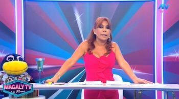 Magaly Medina, vestida con un vestido rosado, permanece de pie tras un escritorio con documentos en un estudio de televisión