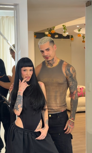 Una mujer de cabello negro largo con flequillo posa frente a un espejo al lado de un hombre con cabello platinado y camiseta sin mangas