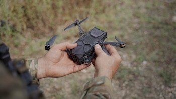Las manos de una persona vestida con uniforme de camuflaje sujetan un pequeño dron negro en un entorno al aire libre