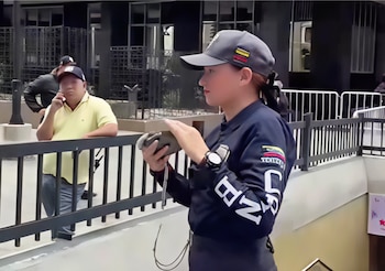 Mujer con gorra gris y uniforme azul oscuro, con emblemas y bandera de Venezuela, sostiene un objeto gris. Un hombre con polo amarillo en segundo plano