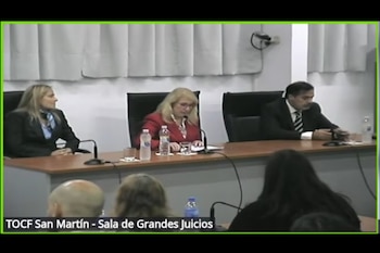 Dos juezas y un juez sentados en una larga mesa de madera con micrófonos y botellas de agua en un tribunal, con público difuminado al frente