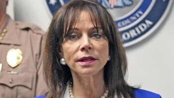 Una mujer de mediana edad con cabello oscuro, collar y pendientes de perlas, con un uniforme y un sello de "County Florida" al fondo