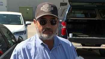 Hombre con barba gris, gafas de sol y gorra negra Harley-Davidson. Viste camisa azul de rayas. Fondo borroso con vehículos y un edificio
