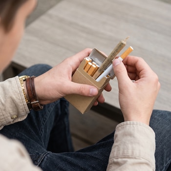 Primer plano de manos sacando un cigarrillo de tabaco y un porro de cannabis de una cajetilla; la persona lleva pulsera de cuero, reloj dorado y ropa beige y vaquera.