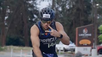 Hombre ciclista con casco, gafas de sol, camiseta negra con número 1105, mochila de hidratación. Saluda con señal de paz. Fondo arbolado