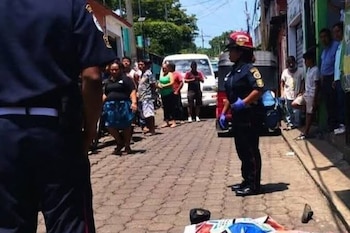 Calle de adoquines con un oficial de policía de uniforme azul y casco rojo, una persona en el suelo cubierta, otro oficial de espalda y varios civiles