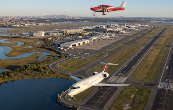 Vista aérea del aeropuerto JFK de Nueva York con un avión de pasajeros blanco y un pequeño avión rojo en vuelo; la ciudad se ve al fondo