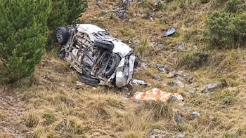 Un vehículo blanco volcado y deformado sobre una pendiente con pasto seco y rocas, junto a una manta colocada en la tierra