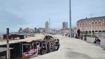 Agentes de la Prefectura Naval Argentina entre puestos de feriantes en la costa de Mar del Plata. Personas observan la escena. Edificios y el mar al fondo bajo cielo nublado