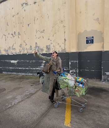 Mujer con abrigo de piel y gafas de sol, bolso y teléfono. Empuja un carro de compras lleno de bolsas. Fondo de pared con pintura descascarada y cartel de prioridad